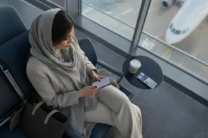 کوچ زن ایرانی در سالن انتظار فرودگاه با موبایل در دست و حالتی آرام پس از منظم شدن رزرو جلسات